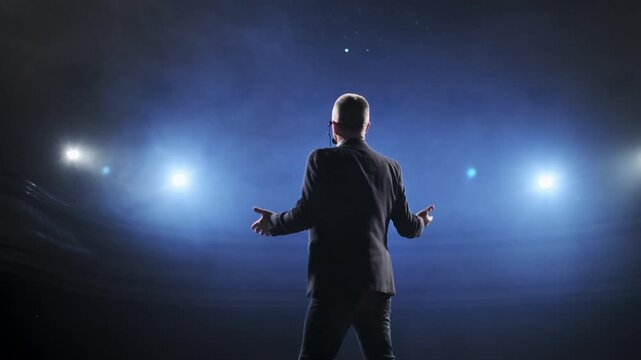 Senior male motivational speaker passionately gestures while delivering an inspiring speech on stage, wearing a headset microphone. Vibrant stage lights illuminate the scene, creating a dynamic and