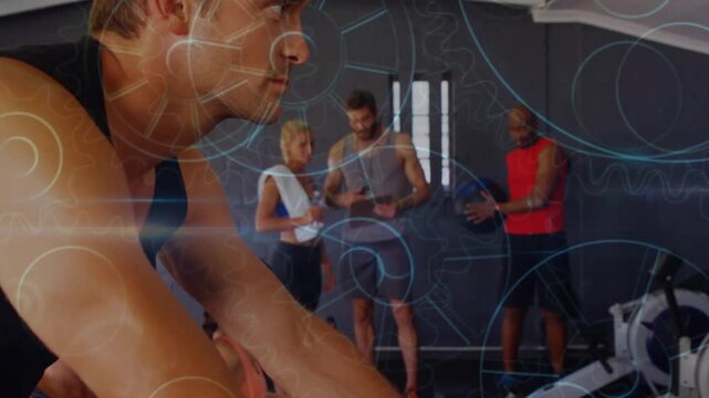 Man biking smiling, turning as camera panning revealing gym HUD, pair reviewing tablet, tracking