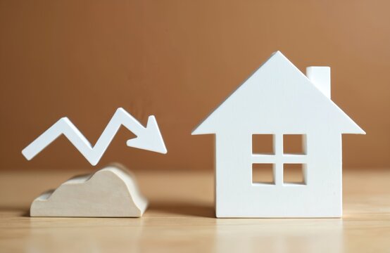 White house model near arrow graph going down on wood surface. Represents real estate market decline, falling property values, or economic recession impacting home ownership.
