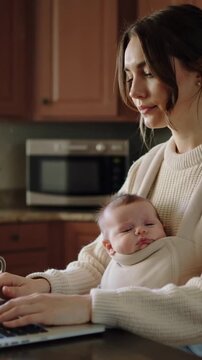 Vertical video: Starting work mom in cream top typing on laptop holding baby in carrier in kitchen