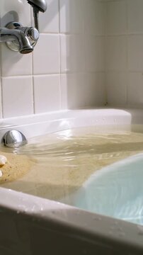 Vertical video: Opening chrome faucet running into tub, filling near sand mound and three seashells