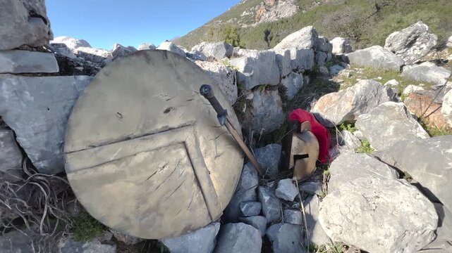 Ancient Greek Spartan helmet with red plume, sword and shield resting against stone ruins outdoors