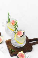 Fototapeta premium Two lemon cocktails with fig rosemary and ice served in glasses on wooden board with fruit and herb decoration