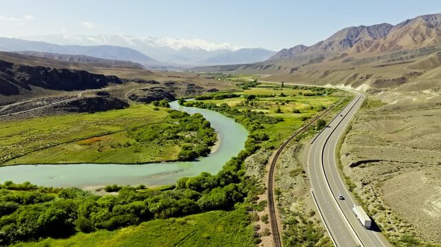 Vast valley features a winding Chu river, modern highway, and railway tracks. Rugged mountains and snow-capped peaks create a tranquil backdrop in daytime.