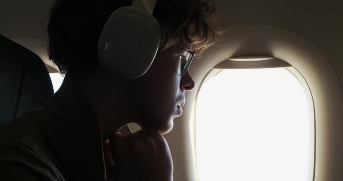 A pensive young person wearing headphones and glasses looks out an airplane window