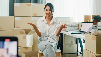 Youth Asia lady owner small business online shop sit front of phone look at camera live streaming talk to customer order promotion with full carton box at home. Shipping and delivery concept. © tirachard