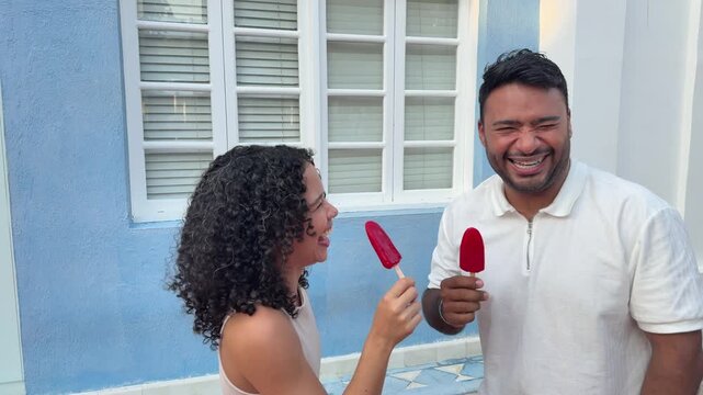 Young biracial couple having a fun date eating popsicles together and laughing in front of a house