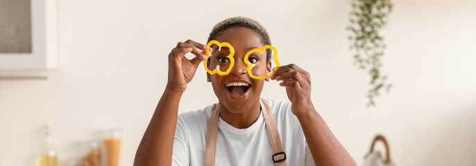 A woman stands in a bright kitchen, smiling widely as she holds yellow bell pepper slices in both hands. She appears to enjoy cooking and having fun with food preparation. © Prostock-studio