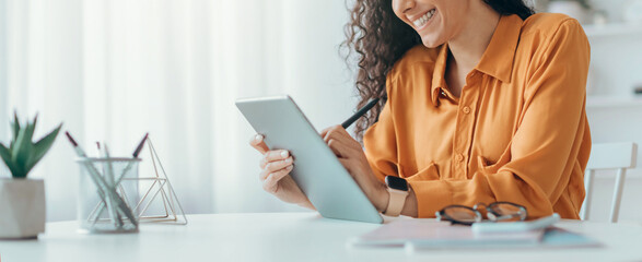 A woman sits at a desk, smiling as she writes on a tablet. She is focused and engaged. In the...