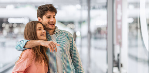 A young couple stands close together, smiling and pointing at items in a shop window. They appear happy and engaged in their shopping experience. The setting is a busy mall during the day. © Prostock-studio