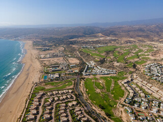 Aerial drone view of the beautiful Taghazout Bay coastline featuring luxury resorts, sandy beaches, lush green golf courses, and the Atlantic Ocean in Agadir, Morocco.