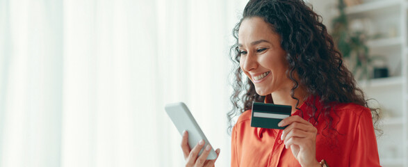 A woman is smiling as she holds a smartphone in one hand and a credit card in the other. She is...