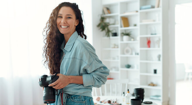 A woman smiles while standing in a bright room. She holds a camera in her hands. The setting includes shelves and items on a table. This scene shows a moment of joy and creativity.