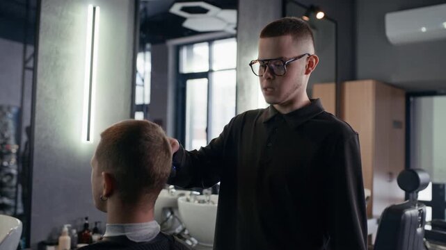 Medium shot of young Caucasian barber chatting with client while trimming his hair during workday in salon