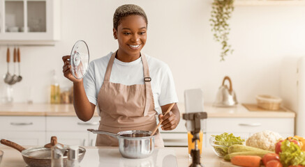 A person stands in a kitchen, holding a lid and stirring a pot on the stove. Fresh vegetables are on the counter. The smartphone is set up for recording a cooking session. © Prostock-studio