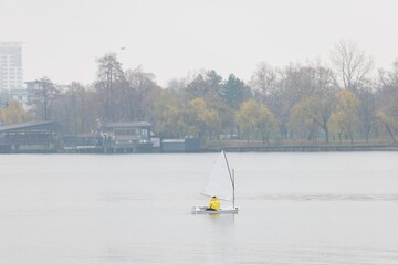A small sailboat glides across a serene lake as a solitary sailor, dressed in a bright yellow raincoat, enjoys the quiet atmosphere. Soft clouds hang overhead, adding a magical touch