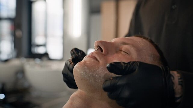 Close up of anonymous barber using cream while massaging face of man who sitting in barbershop chair with closed eyes