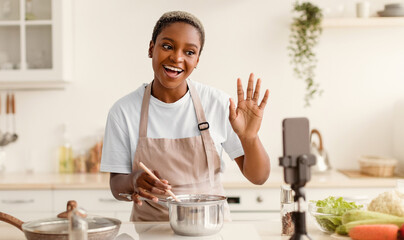 A woman stands in a kitchen with a pot on the stove. She waves to her audience while preparing a meal. Fresh vegetables are on the counter, and she appears engaged with her viewers. © Prostock-studio