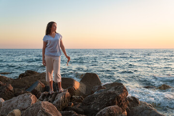 A young woman stands on the rocky coast holding her sunglasses, looking at the horizon during a beautiful summer sunset, symbolizing freedom and inspiration.