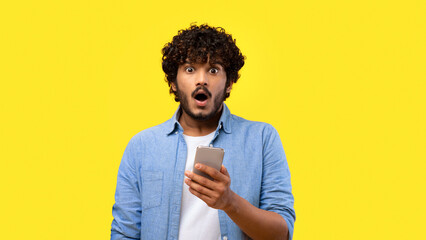 A man stands with a shocked expression as he looks at his smartphone. The background is bright yellow, creating a strong contrast with his casual blue shirt. © Prostock-studio