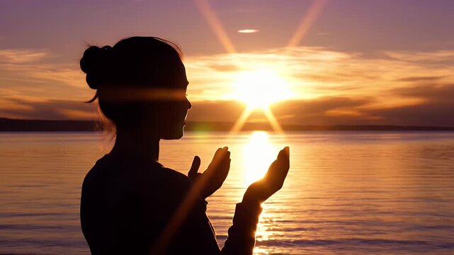 Woman Silhouette Worshiping at Sunset - Silhouette of a woman with open hands and palms facing up in worship against a sunset sky and water.