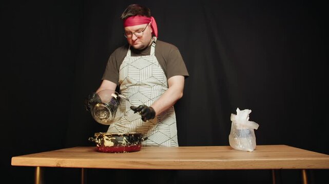 Caucasian man adding raisin to metal pot with batter on wooden table. Chef wearing apron and red headband, black gloves pouring with spoon from glass bowl on black background. Kitchen worker in bakery
