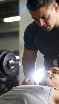 Vertical video: Spotting trainer reacting to lifter lowering dumbbells and pressing on bench in gym