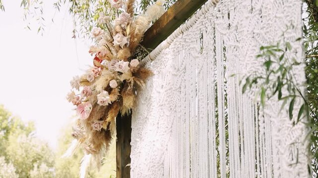 Closeup floral cluster on wedding arch post macrame fringe and pampas accents. Pale pink roses, gypsophila