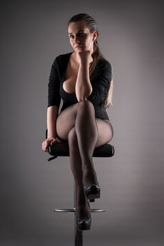 Confident curvy woman in black bodysuit and sheer pantyhose sitting on bar stool with crossed legs