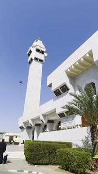 A bright, sunny view of Masjid Aisha (Taneem Mosque) in Makkah, Saudi Arabia. This iconic Miqat site is shown with its unique minaret and modern white architecture as pilgrims prepare for Umrah.