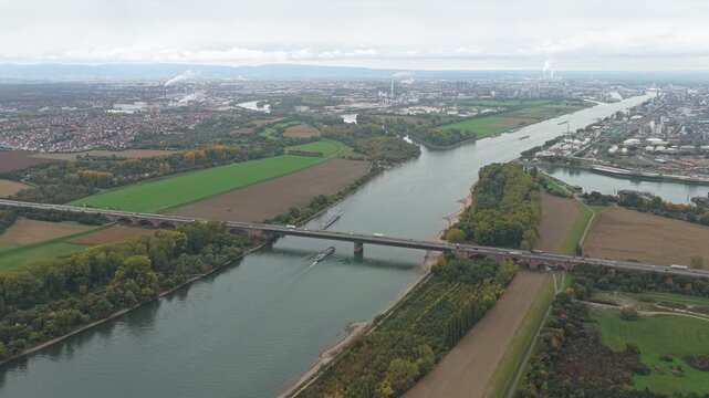 Panoramic aerial view flying over the rhine river, with barges navigating and traffic on a highway bridge, approaching a large industrial complex