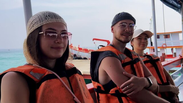 Group of young tourists happily smiles at camera sitting in boat wearing orange life vests before sea trip starts.
