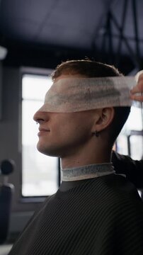 Vertical shot of male client sitting in chair and anonymous barber using hairdressing collar to protect clients eyes during haircutting