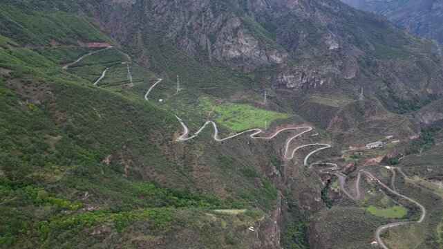 Stunning aerial view of a serpentine road zig-zagging up a steep mountain slope. The winding path cuts through a deep, green gorge, showcasing a dramatic and rugged landscape.

