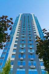 Obraz premium Low angle view of a modern high-rise residential building with glass facade and blue sky background through green tree leaves