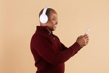 Side view of mature black man using smartphone while wearing wireless headphones on beige studio background. Concept of streaming music, podcast listening and mobile audio entertainment