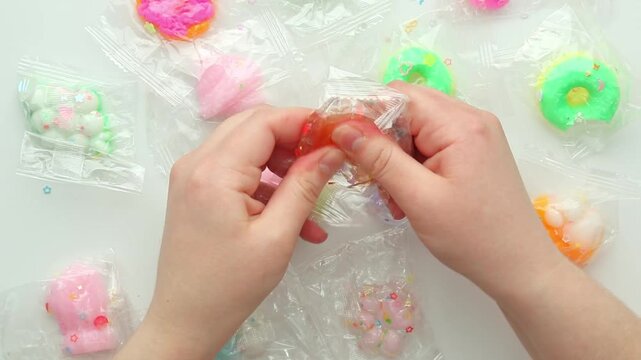 Top view: hands unpack and squeeze a transparent squishy toy, pressing beads inside, then kneading and stretching it over a white table with fidgets, creating satisfying stress-relief motion.
