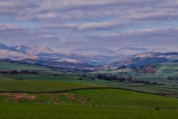 Fototapeta premium Rolling green farmland with stone walls and distant lake and mountains under dramatic cloud cover