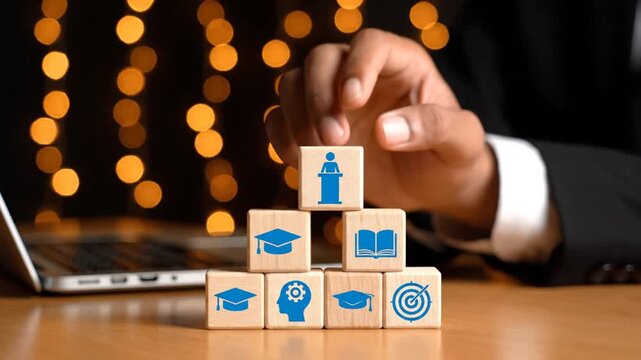 A person builds a pyramid of wooden blocks with business and education icons on a desk next to a laptop in a dark room.