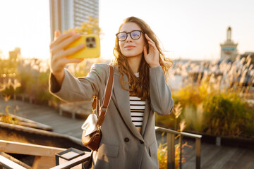 A beautiful woman stands on a rooftop and takes a selfie on her phone at sunset. A young woman enjoys a sunny day and blogs outdoors. Concept of blogging, style, fashion, and technology. © maxbelchenko