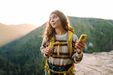 Beautiful woman with phone stands on rocky outcrop at sunset. Hiker with phone and backpack feels free and enjoys mountain scenery while hiking. Concept of nature, technology, and adventure. © maxbelchenko