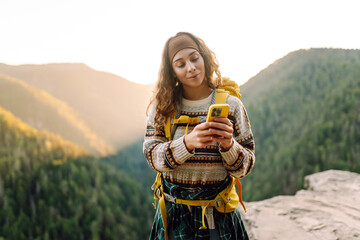 Beautiful woman with phone stands on rocky outcrop at sunset. Hiker with phone and backpack feels free and enjoys mountain scenery while hiking. Concept of nature, technology, and adventure. © maxbelchenko