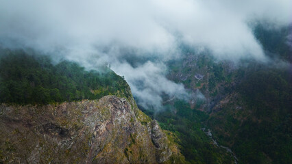The view of Miradouro Eira do Serrado in Madeira, Portugal. A drone flies above the mountains and trees, with clouds filling the valleys below. Madeira aerial view