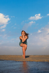 Naklejka premium Woman stands on one leg with the other wrapped, arms crossed, practicing yoga on the beach at sunset.