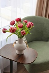 Naklejka premium A beautiful bouquet of pink double tulips in a white ceramic vase on a wooden side table by a green armchair. Concept of cozy spring home decor.