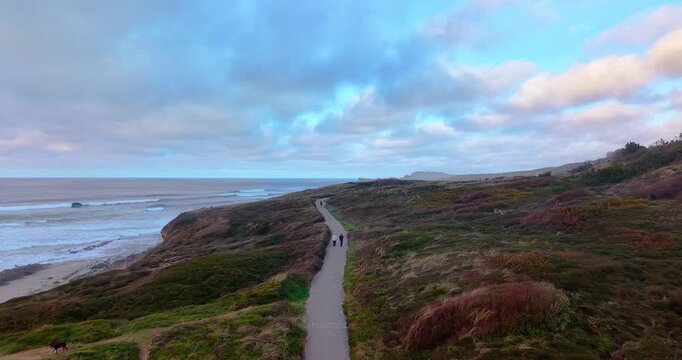 Aerial shot of tourists enjoying a hike on a paved trail through the lush green cliffs of Liencres Dunes Natural Park. The image features the rugged coastline of Cantabrian Sea.
