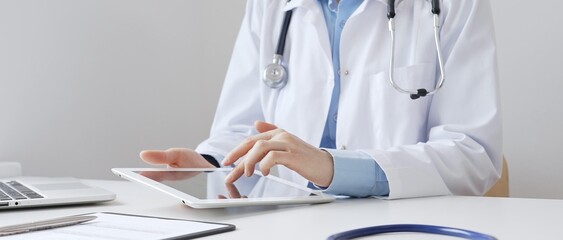 Female doctor wearing white medical coat and stethoscope using digital tablet with laptop and...