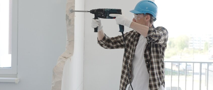 Man construction worker demolishing a wall with a hammer drill in protective gear, creating construction debris on the surrounding floor surface. Renovation concept