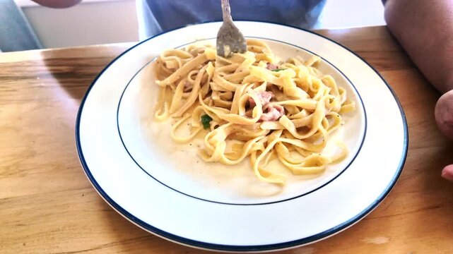 Close-up view of a person twirling and eating a forkful of creamy carbonara pasta from a white plate on a wooden table at lunchtime