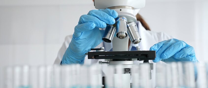Female scientist wearing protective equipment checking microscope, analyzing blue test tubes in modern laboratory research setting. Medicine, healthcare and science concept
